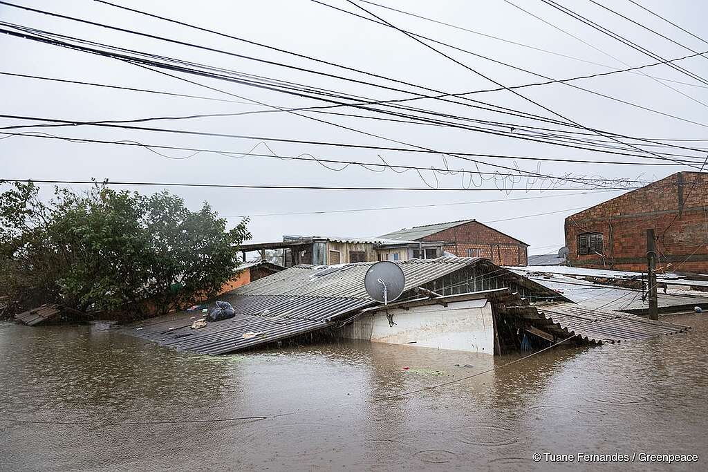 A imagem mostra uma área alagada no bairro Rio Branco, em Canoas, Rio Grande do Sul, afetada por uma grave enchente ocorrida em 2024. A água cobre completamente a casa, mostrando somente um pedaço do telhado e do topo das moradias. No telhado visível, há objetos espalhados, incluindo um saco preto e destroços. A imagem é creditada a Tuane Fernandes.