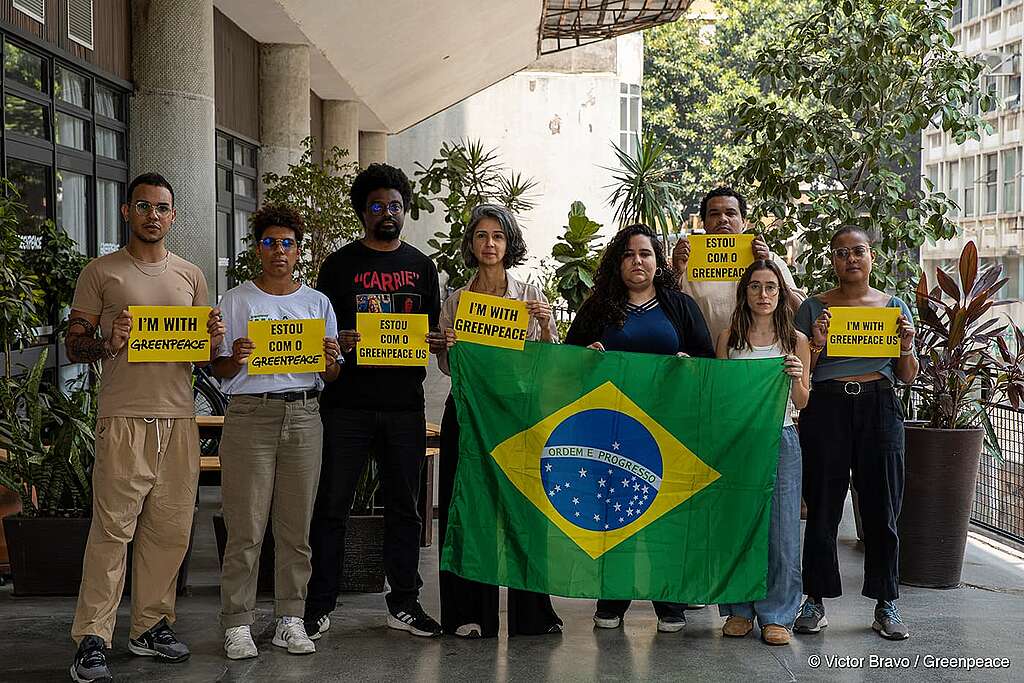 Um grupo de oito pessoas está em pé segurando placas amarelas com mensagens de apoio ao Greenpeace, em inglês e português. Duas delas seguram uma bandeira do Brasil aberta. O cenário é urbano, com plantas e prédios ao fundo. Um grupo de oito pessoas está em pé segurando placas amarelas com mensagens de apoio ao Greenpeace, em inglês e português. Duas delas seguram uma bandeira do Brasil aberta. O cenário é urbano, com plantas e prédios ao fundo.