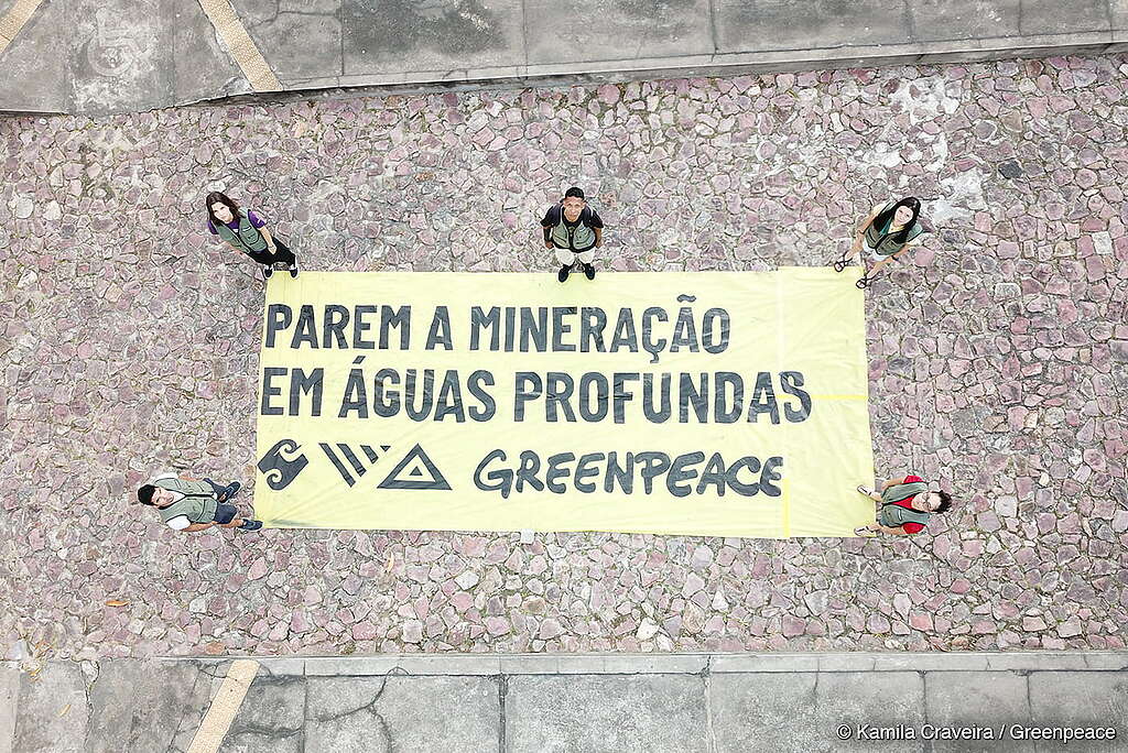 Foto aérea mostra seis voluntários do Greenpeace em pé ao redor de uma grande faixa amarela estendida sobre um chão de pedras. A faixa traz a mensagem “PAREM A MINERAÇÃO EM ÁGUAS PROFUNDAS – GREENPEACE” em letras pretas, acompanhada de símbolos gráficos. A ação acontece em ambiente urbano, com calçamento de pedras e áreas de concreto ao redor.