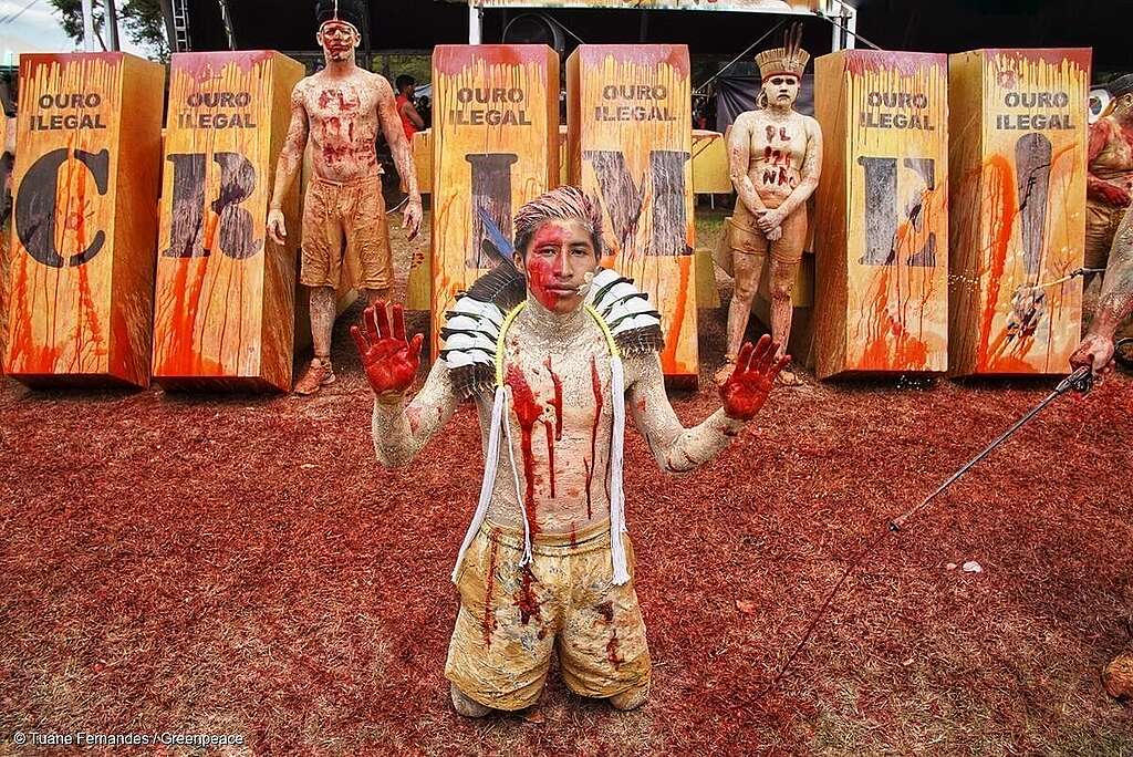 Pessoas indígenas sujas de lama e sangue cenográfico, representando todas as mortes, violências e sofrimento causados pelo garimpo. Na imagem tem blocos de ouro escrito ''Ouro ilegal: crime!''