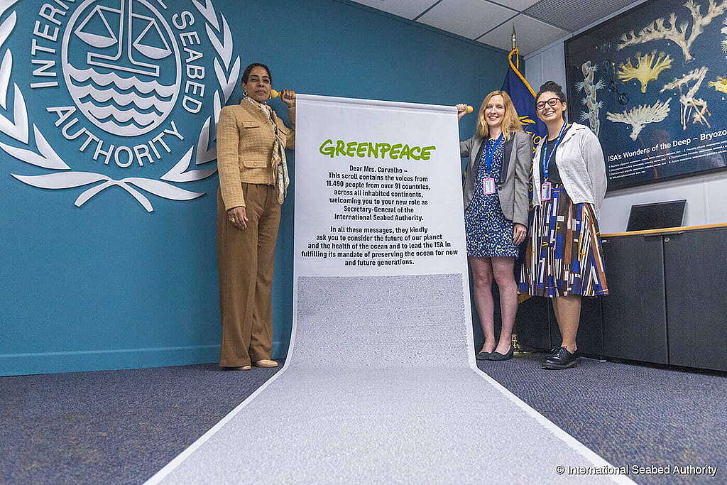 Três mulheres posam sorrindo em frente ao logotipo da Autoridade Internacional dos Fundos Marinhos (ISA), segurando um enorme pergaminho do Greenpeace. O rolo contém mensagens de mais de 11 mil pessoas de 91 países pedindo a proteção dos oceanos. No fundo, há uma parede azul com o emblema da ISA e um painel com organismos marinhos. O pergaminho se desenrola pelo chão, cobrindo boa parte do carpete.