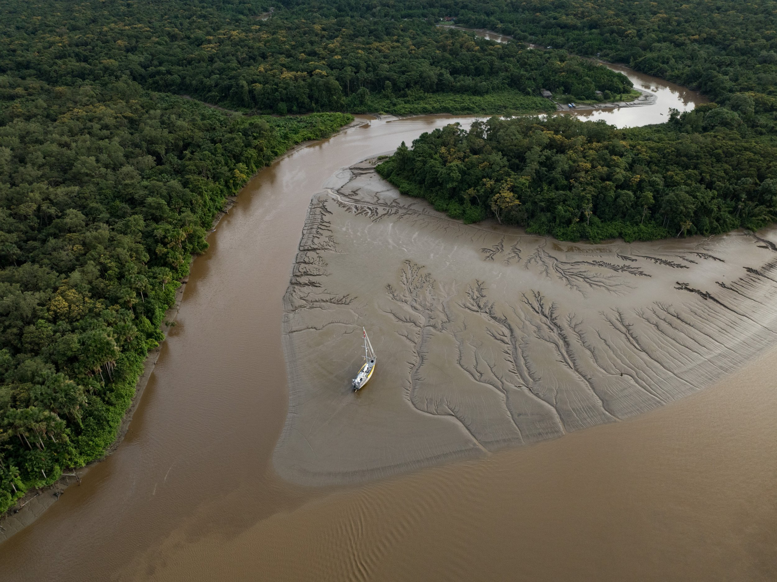 Fotografia aérea de um trecho da costa amazônica durante a maré baixa. No centro da imagem, uma embarcação branca com detalhes amarelos está encalhada em um banco de lama claro, cercado por canais naturais formados pela água. À esquerda e ao fundo, há uma densa floresta verde com árvores altas. Os rios de águas barrentas contornam a vegetação e se dividem em vários braços, compondo uma paisagem sinuosa e rica em biodiversidade.