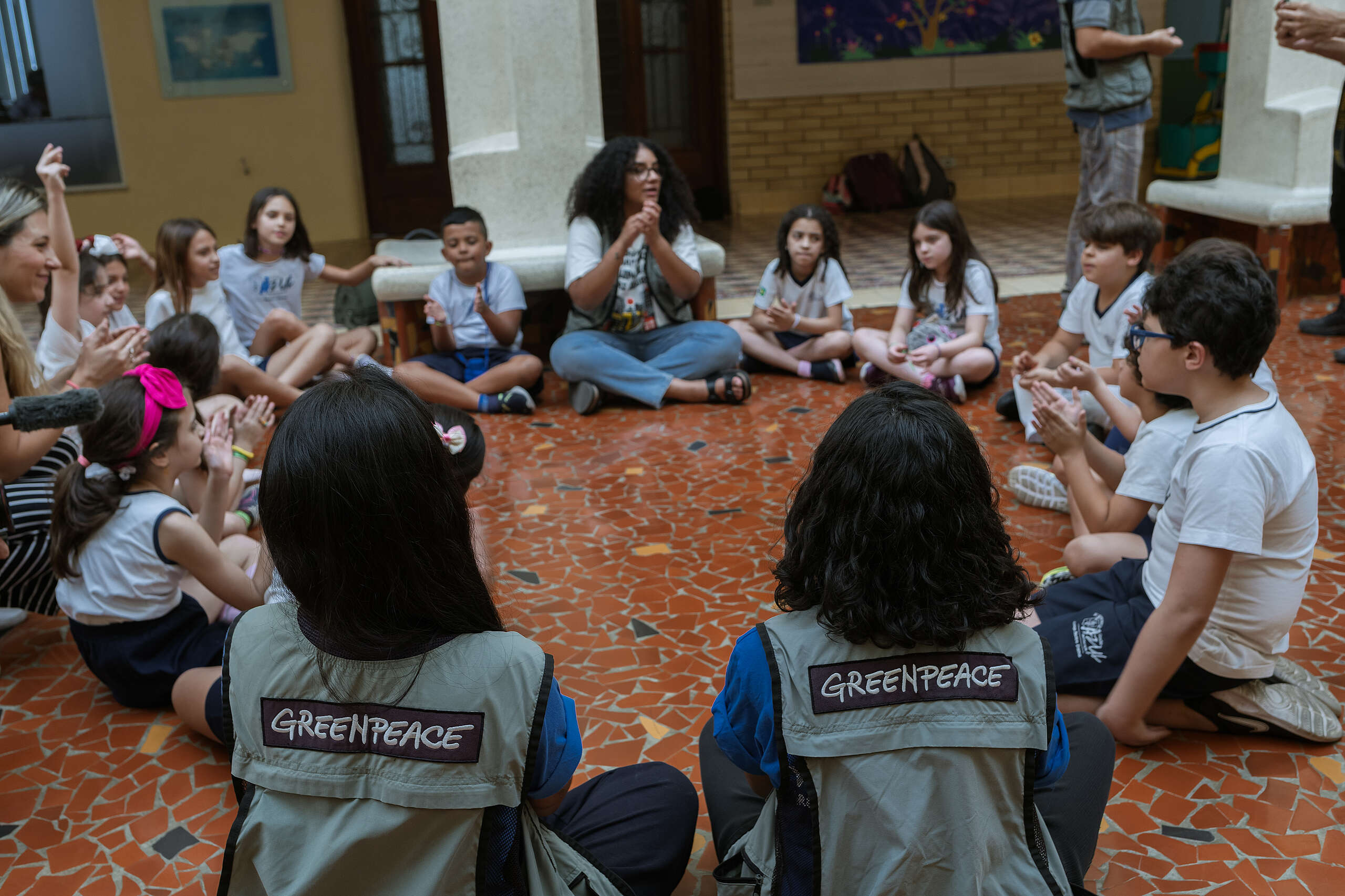 Fotografia de roda de conversa com cerca de 20 pessoas. Na imagem, há três voluntárias com o colete verde-escura escrito Greenpeace nas costas e algumas crianças em uniforme escolar sentadas interagindo com a explicação sobre a importância de salvarmos a Amazônia do desmatamento.