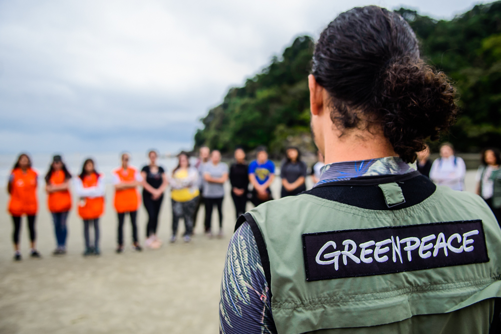 Fotografia com um voluntário do Greenpeace em primeiro plano. Ele está localizado nas areias de uma praia enquanto dá instruções sobre a importância da preservação ambiental para um grupo de alunos que o observa atentamente. O grupo está nos preparativos para iniciar uma atividade de limpeza nesta praia.