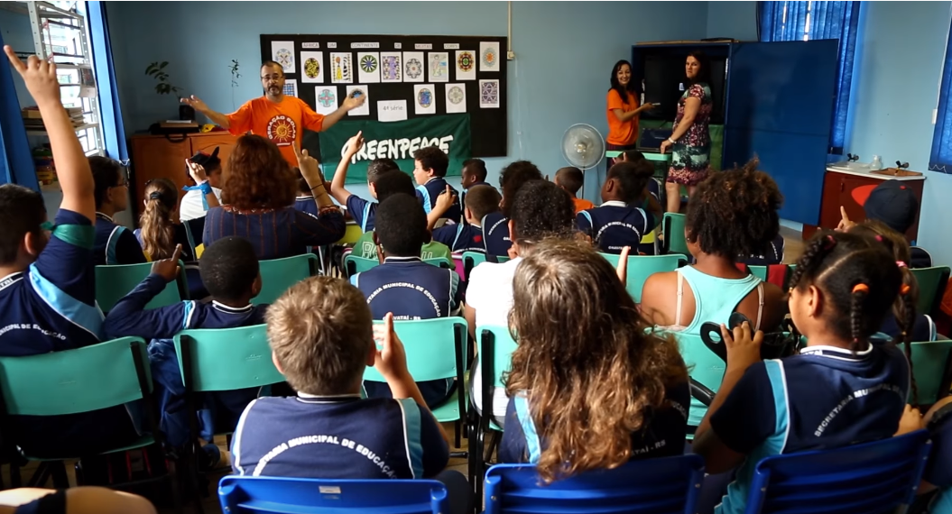 Fotografia com dezenas de estudantes sentados em cadeiras, na sala de aula. Na frente dos estudantes há um voluntário do Greenpeace fazendo um questionamento, na qual alguns estudantes levantam a mão para responder.