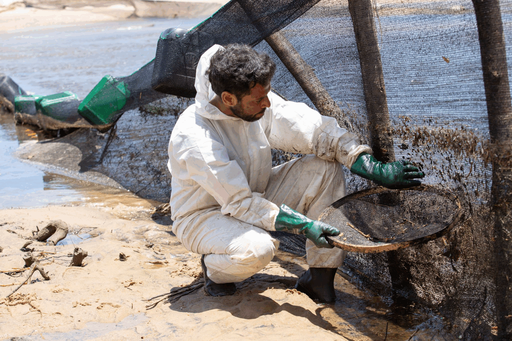 Fotógrafo e morador local, criador do sistema de filtragem com boias no rio Piracanga, atua na limpeza.
Ele veste macacão branco, botas e luvas verdes enquanto ajusta a barreira de contenção.
Na peneira, retira fragmentos de óleo retidos pela rede instalada no manguezal.
A cena mostra o encontro de areia, água e vegetação impactada.