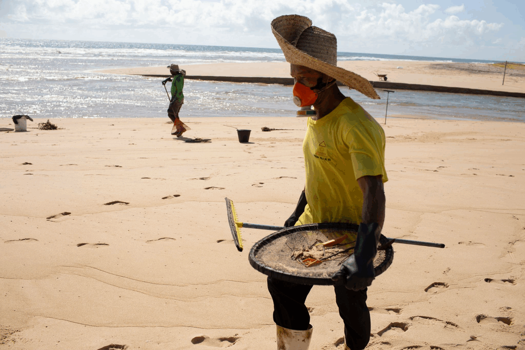 Homem com chapéu de palha, máscara e luvas caminha pela praia segurando peneira com resíduos.
Ele participa da coleta após o derramamento de óleo no Nordeste em 2019.
Ao fundo, outro trabalhador utiliza ferramenta para retirar contaminantes.
O cenário mostra areia clara, mar ao fundo e marcas de passos.