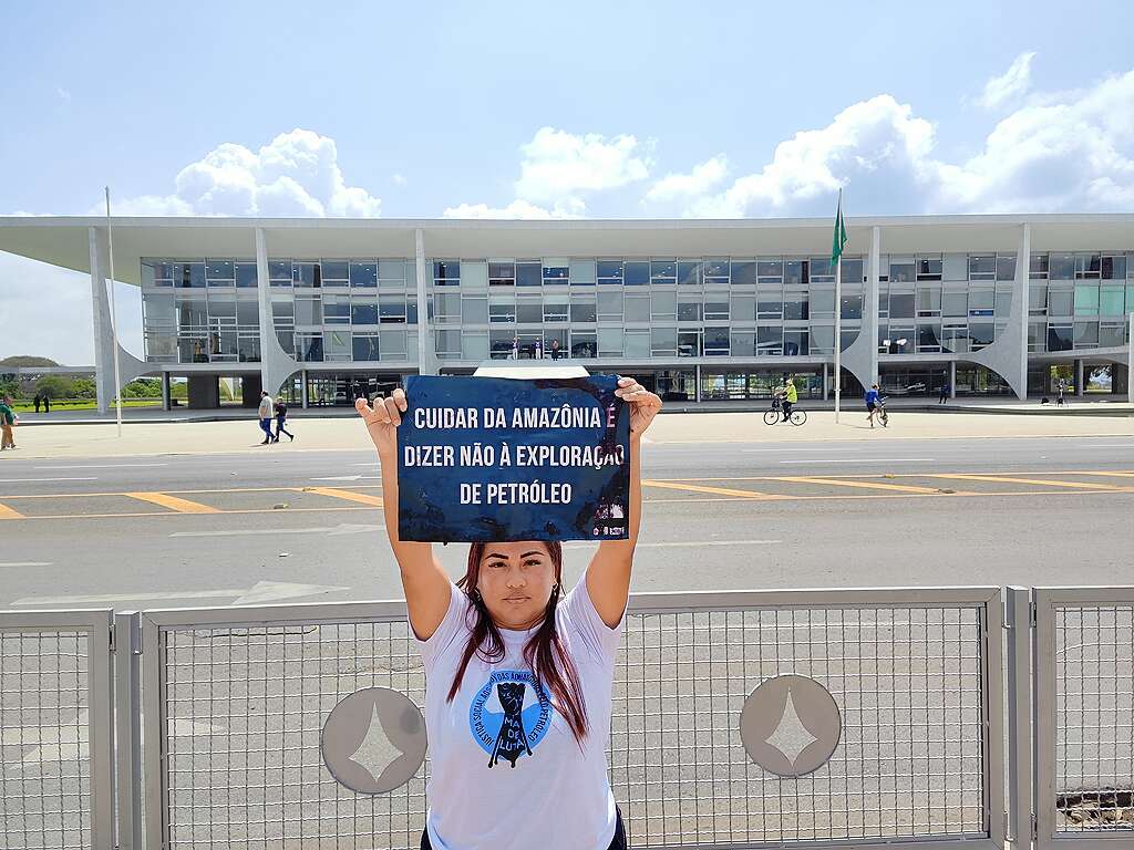 Mulher segura um cartaz em frente ao Palácio do Planalto, em Brasília.
No cartaz, a mensagem: “Cuidar da Amazônia é dizer não à exploração de petróleo”.
Ela veste camiseta branca da campanha Mar de Luta.
O edifício moderno do governo e a bandeira do Brasil aparecem ao fundo.