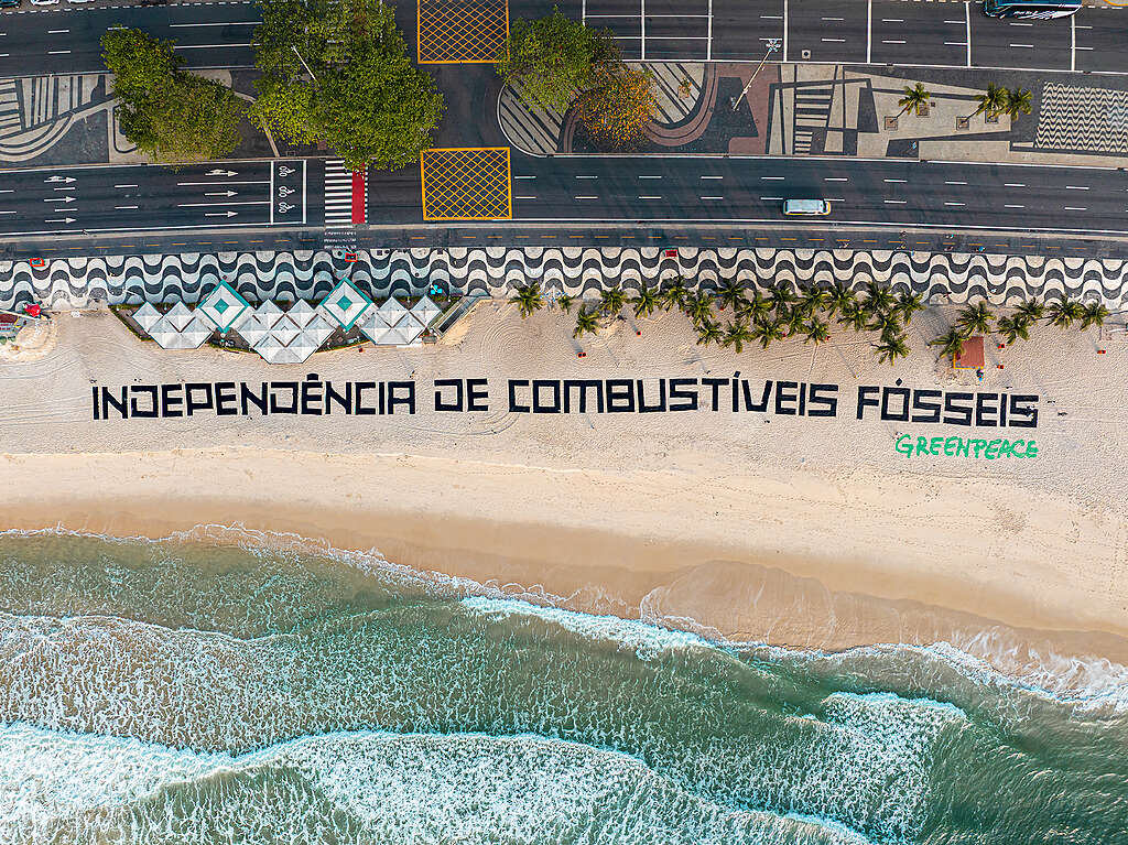 Vista aérea da Praia de Copacabana, no Rio de Janeiro, com grandes letras pretas na areia formando a frase 'INDEPENDÊNCIA DE COMBUSTÍVEIS FÓSSEIS'. O mar ocupa a parte inferior da imagem, enquanto a orla e o calçadão com ondas em preto e branco aparecem acima. Ao lado da frase, em verde, está escrito 'Greenpeace'.