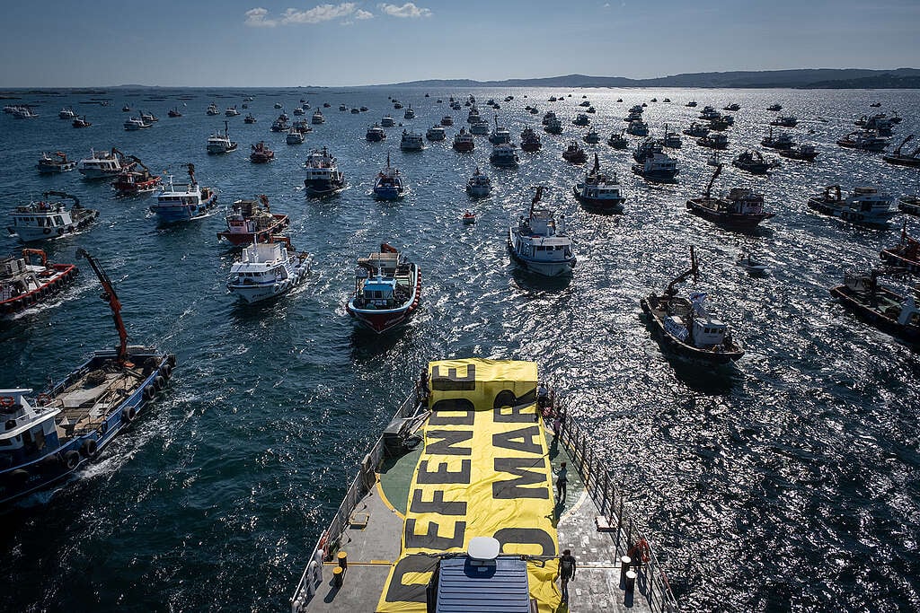 Diversas embarcações ocupam o mar em manifestação. No primeiro plano, o convés de um navio exibe uma grande faixa amarela com a mensagem “Defenda o Mar”.