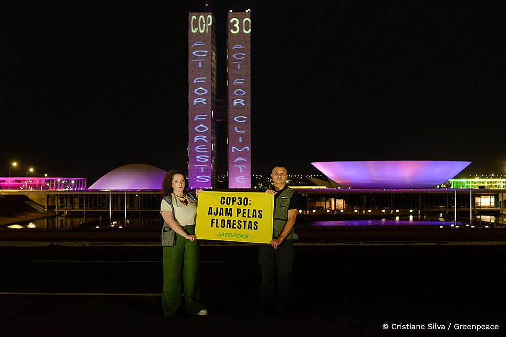 Foto de dois ativistas no Congresso Nacional, em Brasília, segurando um banner com os dizeres “COP30: ajam pelas florestas”. Ao fundo, as duas torres do Congresso exibem uma projeção com as frases “COP: ACT FOR FORESTS” e “COP30: ACT FOR CLIMATE”.