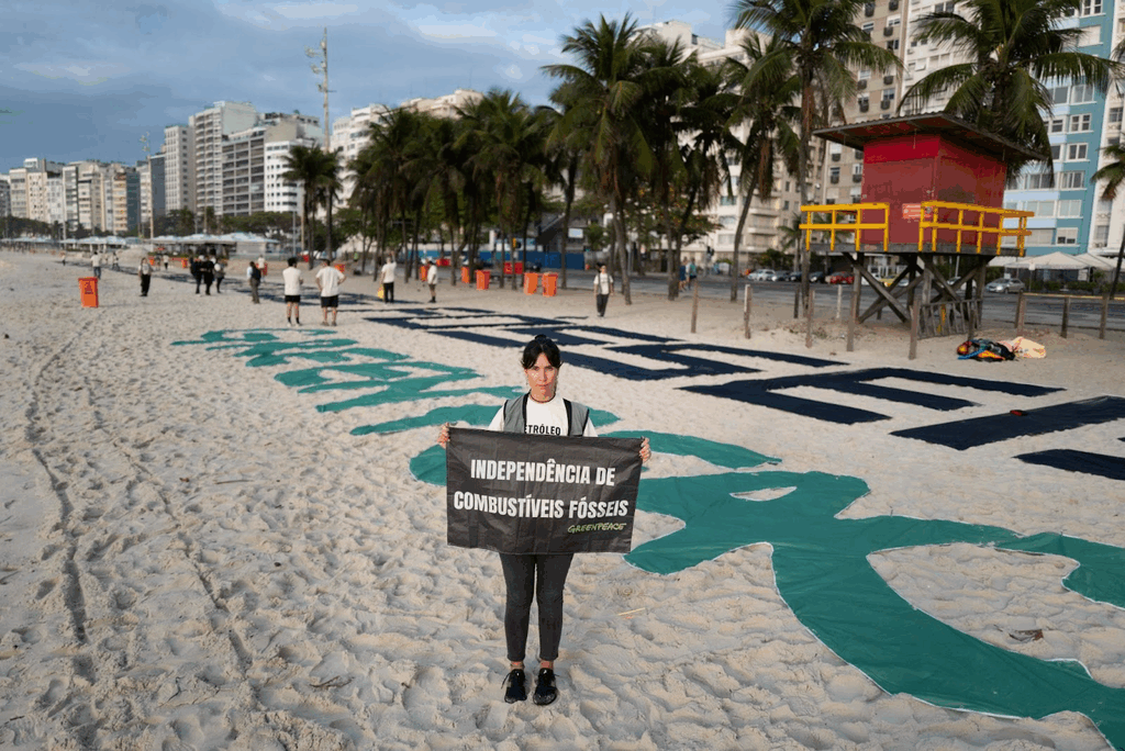 Mulher segura um cartaz preto com os dizeres “INDEPENDÊNCIA DE COMBUSTÍVEIS FÓSSEIS – Greenpeace” em uma praia. Ao fundo, outras pessoas posicionam grandes letras verdes e pretas sobre a areia formando uma mensagem gigante.