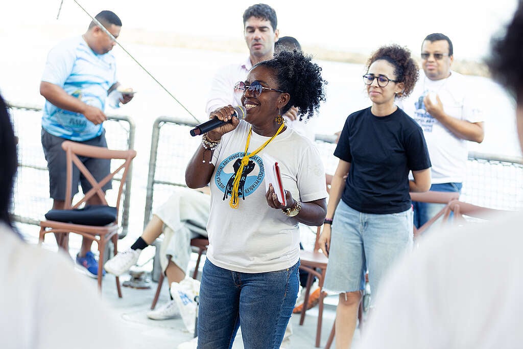 Uma mulher negra, sorrindo, fala ao microfone no convés do navio Rainbow Warrior durante o encontro com lideranças da pesca artesanal. Ela usa óculos escuros, camiseta branca com o símbolo do Movimento Mar de Luta e segura um caderno vermelho. Outras pessoas, sentadas e em pé, acompanham a fala ao fundo. A cena transmite acolhimento, força e participação ativa das comunidades costeiras.