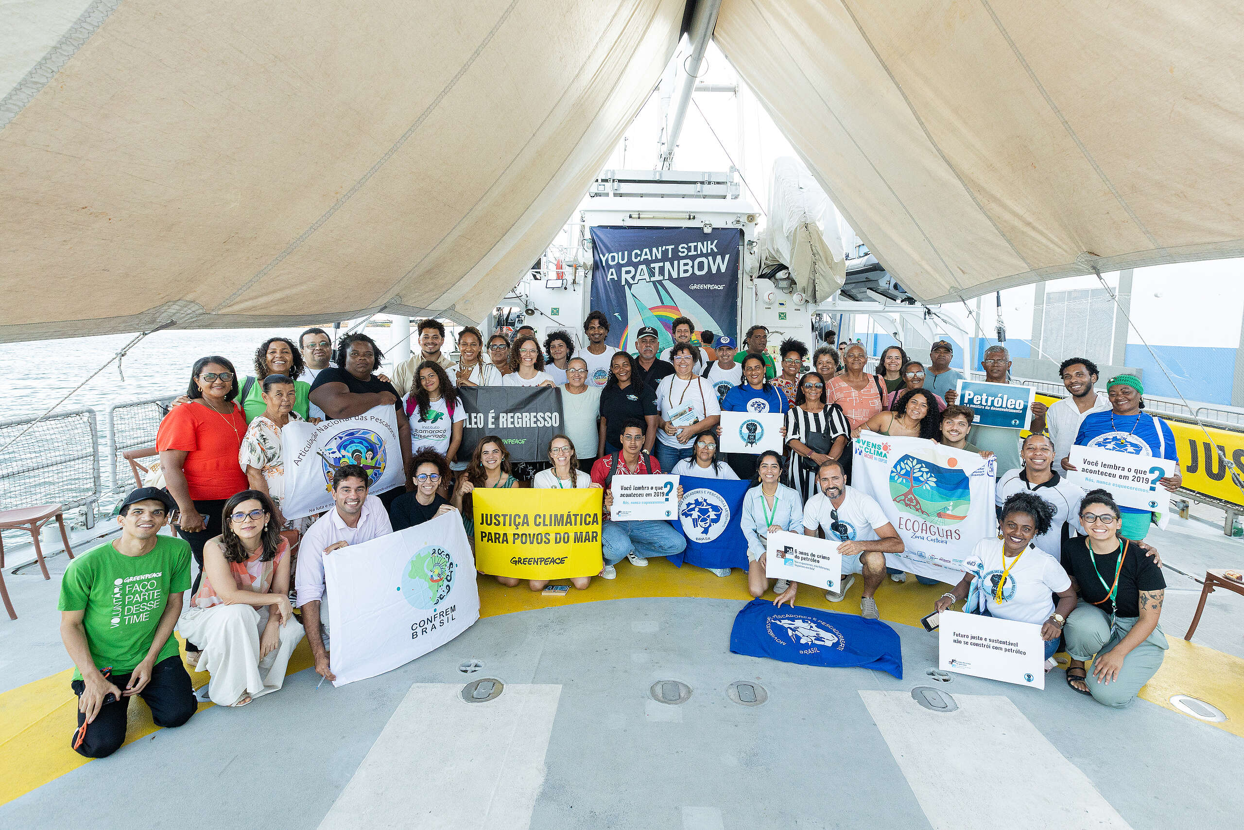 Cerca de cinquenta pessoas, entre marisqueiras, pescadores, lideranças comunitárias, jovens e integrantes do Greenpeace, posam juntas no convés do navio Rainbow Warrior. O grupo segura faixas e cartazes com mensagens sobre justiça climática, defesa dos oceanos e memória do derramamento de óleo de 2019. Todxs sorriem e demonstram união e força coletiva. Ao fundo, aparece a parte traseira do navio com o banner “You Can’t Sink a Rainbow”. A cena transmite diversidade, acolhimento e mobilização dos povos do mar.