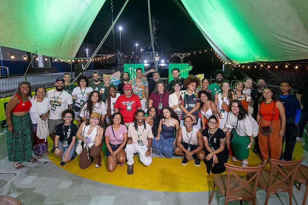 Um grande grupo de pessoas posa reunido no convés de um navio durante a noite, sob uma estrutura de lona iluminada por luzes verdes e cordões de lâmpadas. Há diversidade de idades, corpos e identidades, com integrantes de movimentos sociais, artistas e equipe do Greenpeace. Todas sorriem e demonstram proximidade e celebração coletiva. A cena transmite encontro, afeto e fortalecimento das lutas socioambientais.