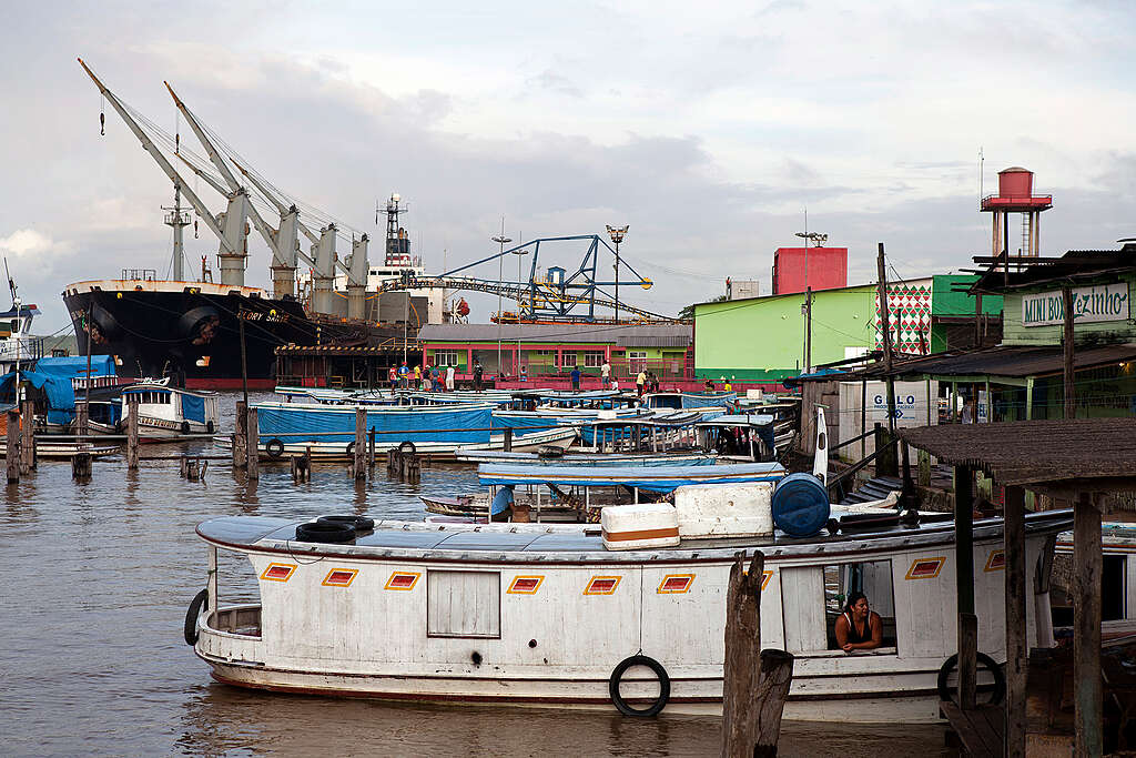 Em um porto amazônico, diversos barcos de madeira estão ancorados próximos a um grande navio de carga ao fundo. As construções coloridas às margens do rio compõem a paisagem urbana ribeirinha. Em primeiro plano, uma mulher observa o movimento de dentro de um dos barcos. A cena retrata o cotidiano das comunidades que vivem e trabalham às margens da água.