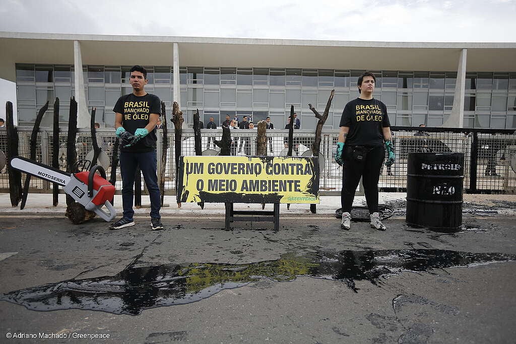 Dois ativistas do Greenpeace vestindo camisetas pretas com a frase “Brasil manchado de óleo” participam de um protesto em frente ao Palácio do Planalto. Eles estão ao lado de cercas queimadas, um tambor preto marcado como “petróleo” e uma motosserra cenográfica. À frente, um cartaz amarelo diz “Um governo contra o meio ambiente”. Manchas escuras simulando óleo espalham-se pelo chão, simbolizando destruição e descaso ambiental.