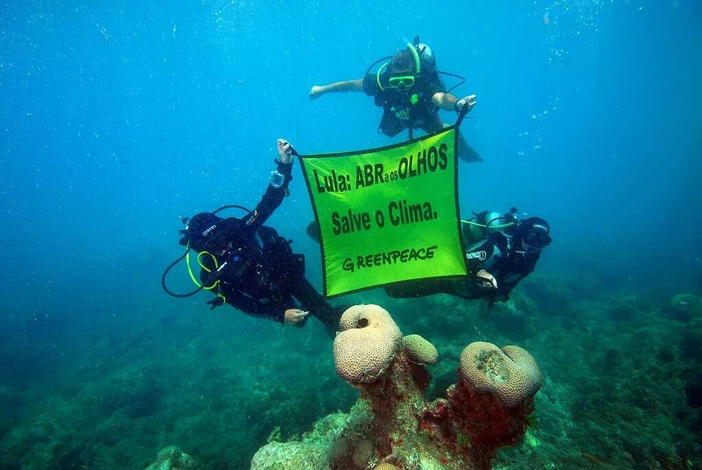 Três mergulhadores equipados com cilindros de ar e máscaras seguram um banner verde debaixo d’água. A mensagem no tecido diz “Lula: abra os olhos. Salve o clima. Greenpeace.” À frente deles, corais aparecem no fundo do mar. A cena registra um protesto submarino em defesa do clima e da proteção dos oceanos.