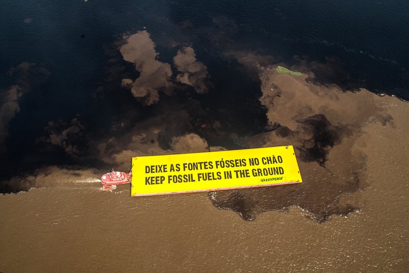 Vista aérea de uma grande mancha de óleo escuro espalhada sobre a superfície do mar. Ao lado da área poluída, um barco vermelho conduz um imenso banner amarelo com a mensagem “Deixe as fontes fósseis no chão – Keep fossil fuels in the ground”. A cena evidencia a gravidade de um derramamento de petróleo e o chamado urgente por justiça climática.