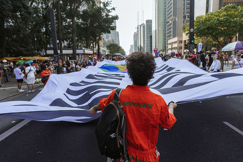 Pessoa com macacão laranja do Greenpeace segura uma grande faixa em manifestação urbana.
Ato ocorre em avenida cheia, com prédios altos e pessoas reunidas ao redor.