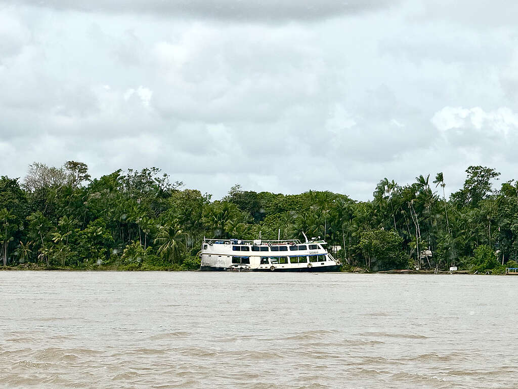 Barco branco de dois andares parado à margem de um rio largo e barrento, em frente a uma faixa densa de vegetação amazônica. O céu está tomado por nuvens cinzas e baixas, criando uma paisagem úmida e silenciosa.