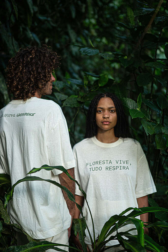 Duas pessoas em meio à floresta usando camisetas claras; uma delas encara a câmera, enquanto a outra está de costas. Na camiseta em destaque, lê-se “Floresta vive, tudo respira”. Nas costas, as logos de Flavia Aranha e Greenpeace Brasil