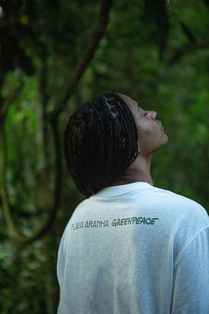 Pessoa em meio à floresta, vista de perfil e de costas, usando camiseta clara com as logos das marcas Flavia Aranha e Greenpeace, enquanto observa a vegetação ao redor.