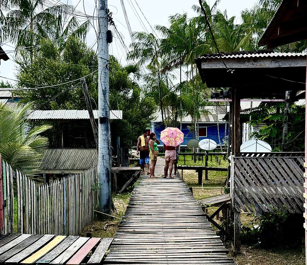 Passarela de madeira estreita atravessa uma comunidade com casas simples, cercas e muitas palmeiras ao redor. Ao fundo, três pessoas conversam sobre a ponte, uma delas segurando um guarda-chuva rosa colorido.