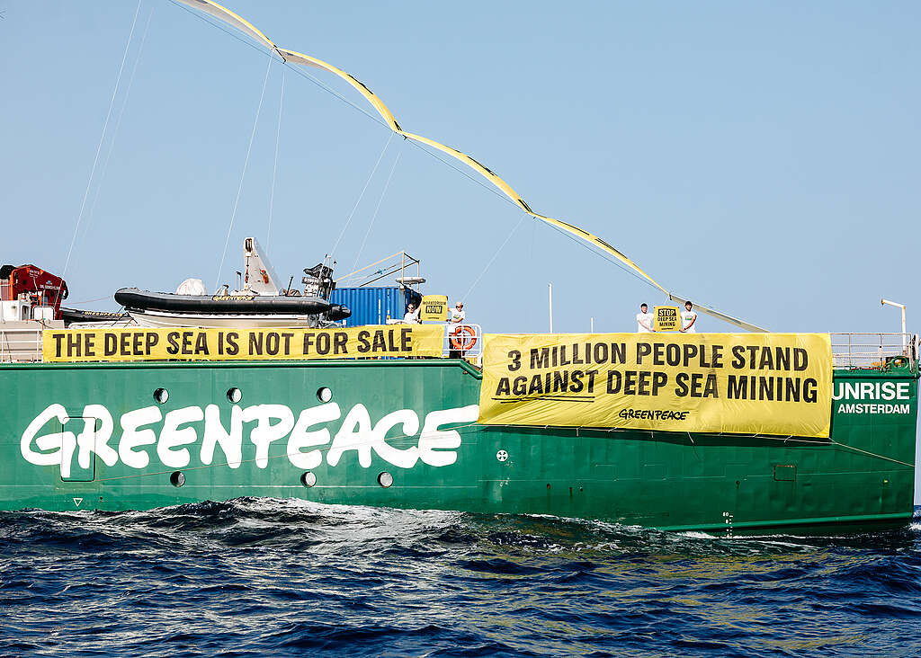 Navio do Greenpeace navega no mar com faixas amarelas de protesto.
Banners afirmam que o mar profundo não está à venda e denunciam a mineração em águas profundas.