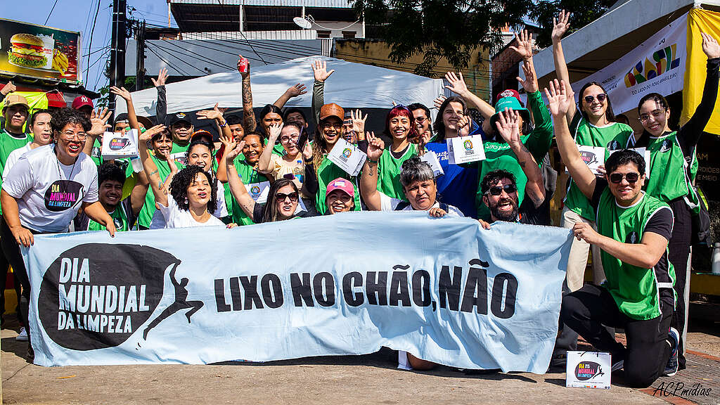 Grupo de participantes posa sorrindo e com os braços erguidos atrás de uma faixa azul com a frase “Lixo no chão, não”, durante uma mobilização do Dia Mundial da Limpeza em área urbana.