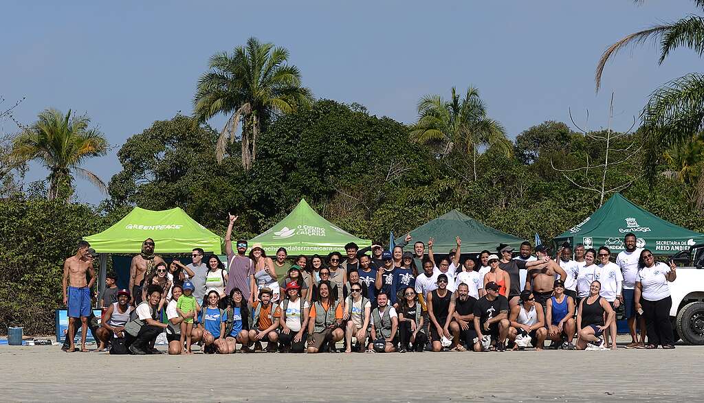 Grupo grande de participantes posa em uma praia diante de tendas de organizações e da vegetação ao fundo, após uma ação coletiva de limpeza e mobilização socioambiental.