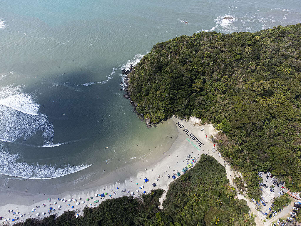 Vista aérea de uma praia cercada por mata verde e mar azul-esverdeado. Na faixa de areia, há uma grande intervenção com a frase “NO PLASTIC”, enquanto pessoas aproveitam a praia e o mar ao redor.