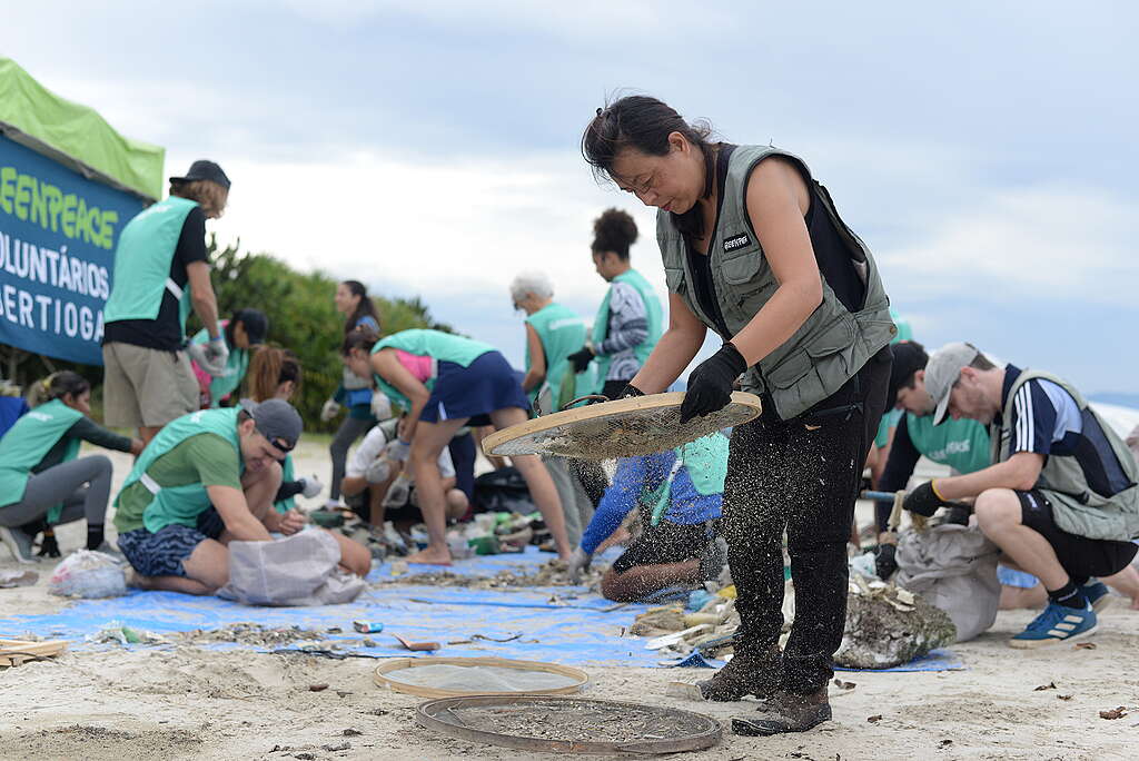 Voluntários participam de uma ação de limpeza na praia. Em primeiro plano, uma mulher peneira areia sobre uma lona azul enquanto outras pessoas ao redor recolhem resíduos com luvas e equipamentos.