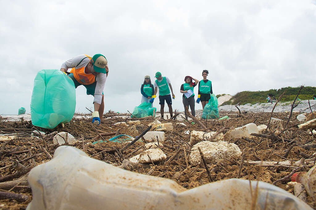 Pessoas recolhem lixo espalhado em uma praia coberta por galhos, plásticos e resíduos. Em primeiro plano, um participante se abaixa com um grande saco verde, enquanto outros voluntários caminham ao fundo.
