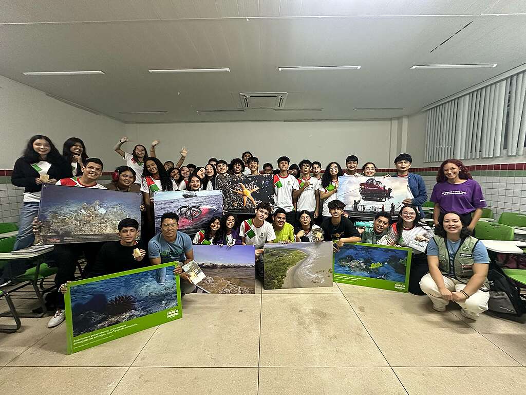 Grupo grande de estudantes e apoiadores posa dentro de uma sala de aula segurando fotografias ampliadas de paisagens marinhas, praias e vida oceânica. Algumas pessoas estão sorrindo e acenando para a câmera.