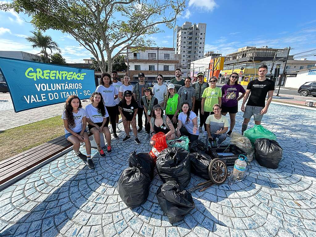 Grupo de voluntários do Greenpeace posa ao ar livre ao lado de um banner com a identificação “Voluntários Vale do Itajaí - SC”. Em frente ao grupo, há vários sacos de lixo recolhido durante uma ação de limpeza.