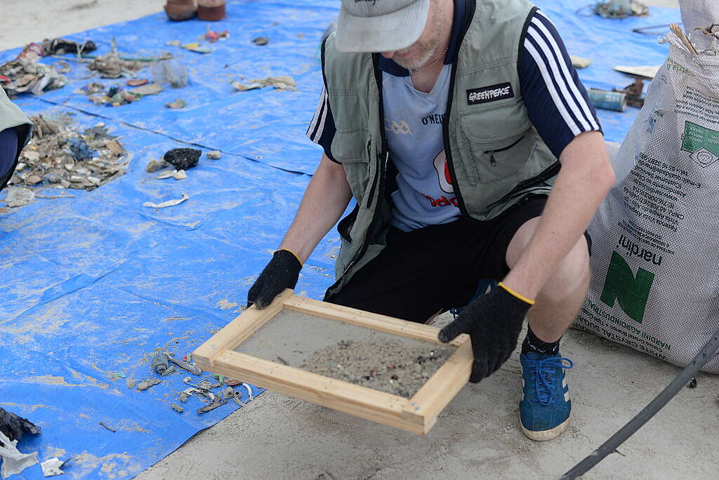 Homem ajoelhado na areia segura uma peneira de madeira cheia de areia fina, durante atividade de triagem de resíduos em uma praia. Ao redor dele, há lona azul, fragmentos de lixo e sacos de coleta.
