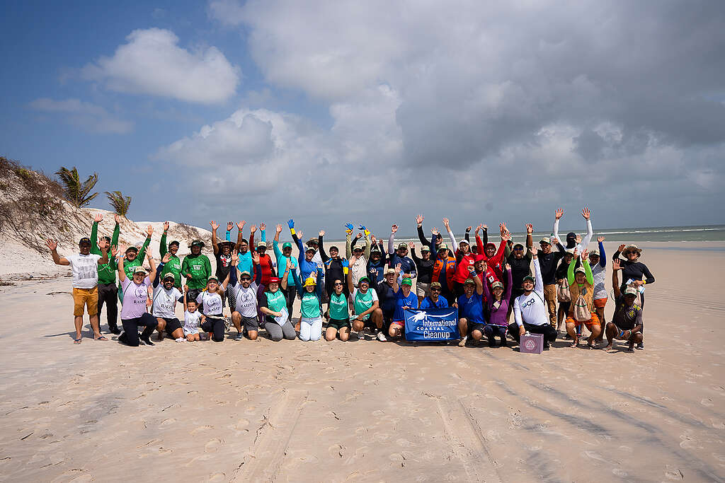 Grupo numeroso de participantes posa em uma praia de areia clara durante uma ação coletiva de limpeza costeira. Todos estão reunidos em frente ao mar, muitos com os braços erguidos, ao redor de uma faixa da International Coastal Cleanup.