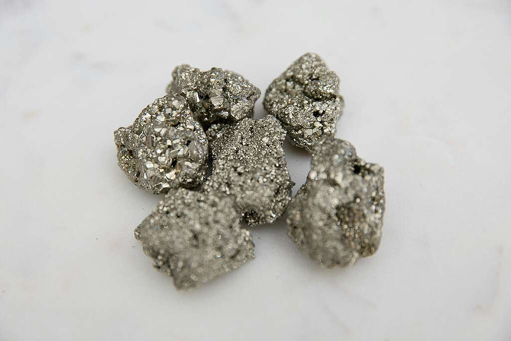 Imagem em close-up mostra pequenos fragmentos minerais met&aacute;licos sobre uma superf&iacute;cie clara. As pedras t&ecirc;m textura irregular, apar&ecirc;ncia prateada e pontos brilhantes que refletem a luz. O fundo est&aacute; desfocado, destacando o volume e o aspecto rugoso dos minerais.