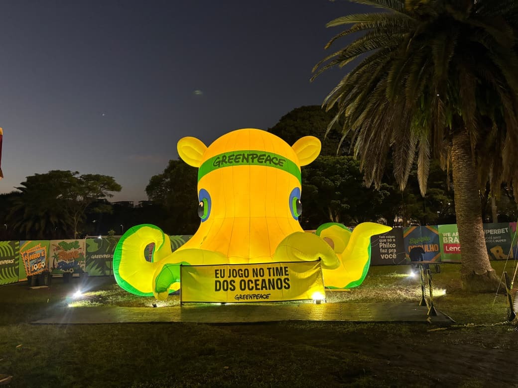 Polvo inflável amarelo e iluminado do Greenpeace instalado em um gramado ao ar livre durante a noite. À frente da estrutura, há uma faixa amarela em destaque com os dizeres "EU JOGO NO TIME DOS OCEANOS".
