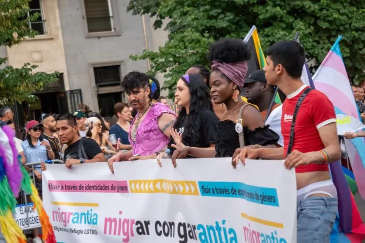 Grupo diverso de pessoas caminha em uma manifesta&ccedil;&atilde;o segurando uma grande faixa branca com os dizeres "migrantia" e "migrar con garant&iacute;a", cercado por bandeiras do orgulho LGBTQIA+ e trans.