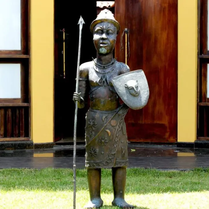 Est&aacute;tua de metal de um guerreiro africano em p&eacute; sobre a grama, segurando uma lan&ccedil;a comprida na m&atilde;o direita e um escudo com a cabe&ccedil;a de um felino na esquerda, posicionada em frente a uma porta de madeira.