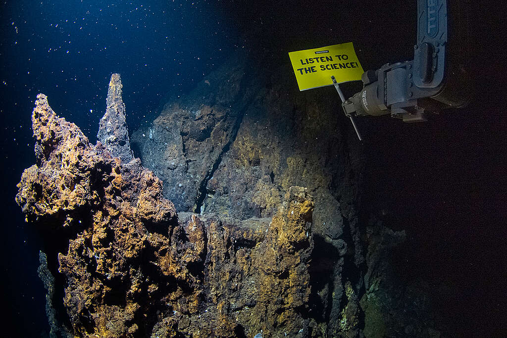 Robô submarino segura uma placa com a frase “Listen to the Science” próximo a formações rochosas no fundo do mar. A imagem reforça o alerta da ciência diante da pressão pelo início da exploração de minerais críticos no oceano profundo.
