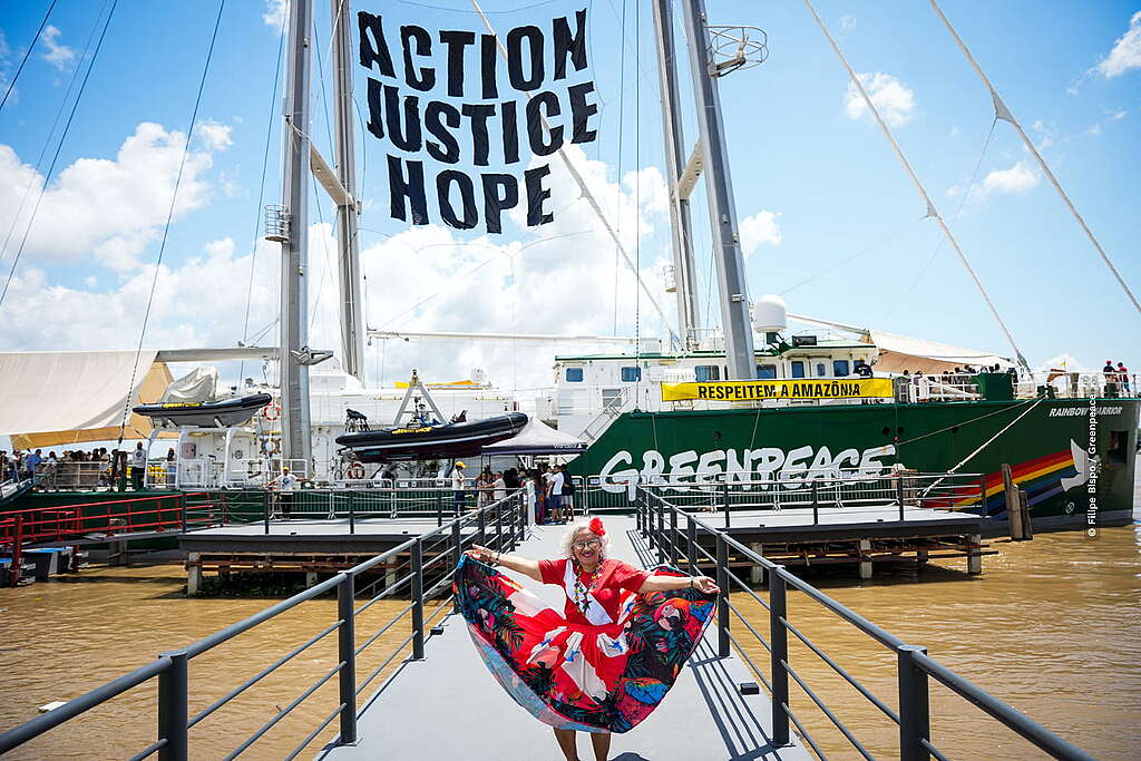 Mulher com vestido vermelho estampado posa sorrindo na passarela de acesso ao navio Rainbow Warrior, do Greenpeace. Ao fundo, o navio verde exibe uma faixa amarela com a frase "AÇÃO. JUSTIÇA. ESPERANÇA." em inglês e outra com a mensagem "RESPEITEM A AMAZÔNIA", em português.