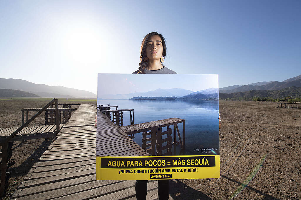 A Greenpeace activist protests at Laguna de Aculeo for urgent and ambitious action on climate in 2019, holding a poster. The image on the poster is of the lake when it was full of water, which contrasts the dry basin in the background.