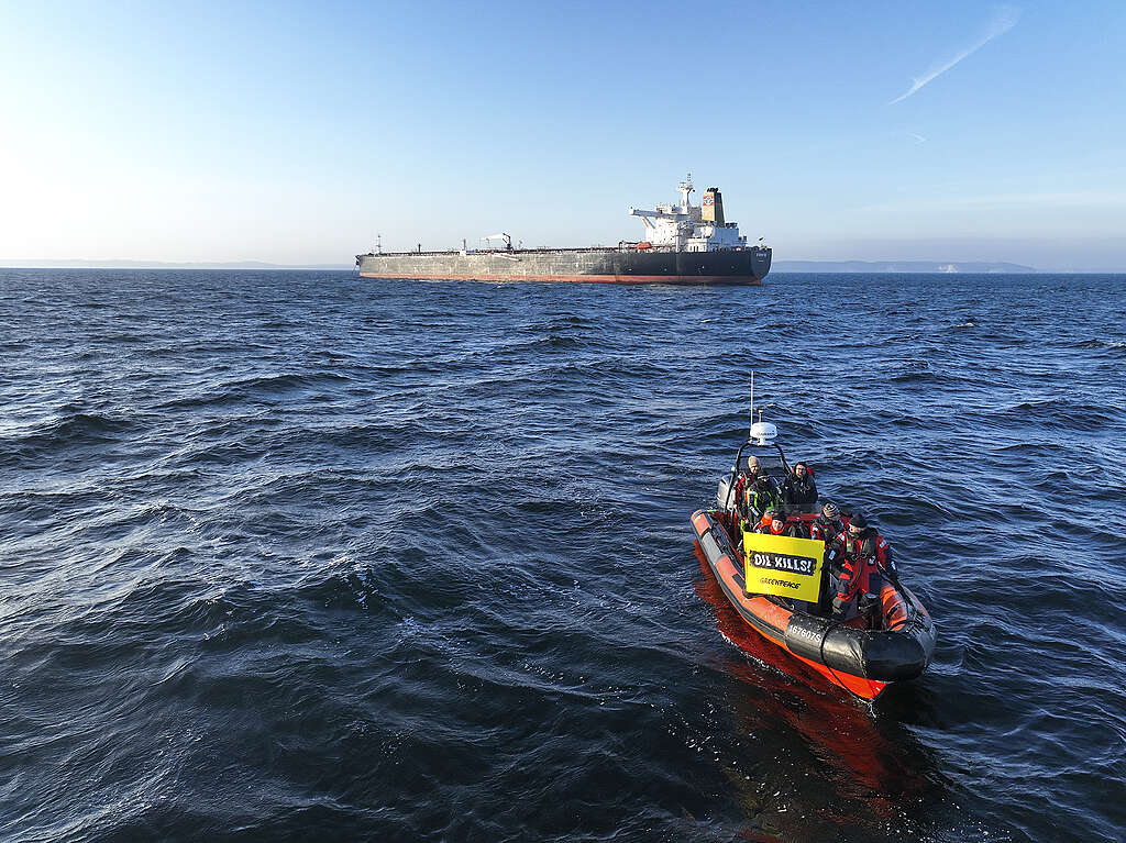 The image shows a group of Greenpeace activists sailing towards a tanker on an inflatable, holding a banner that read "oil kills."