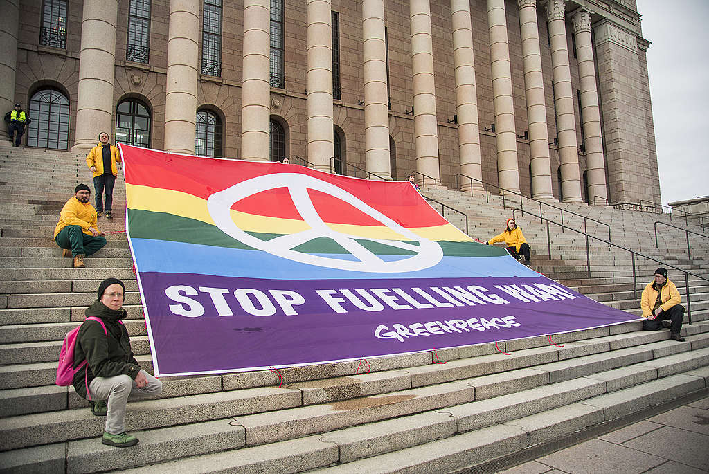 Seitsemän mielenosoittajaa pitävät Eduskuntatalon edustalla Greenpeacen banderollia, jossa on teksti "stop fuelling war".