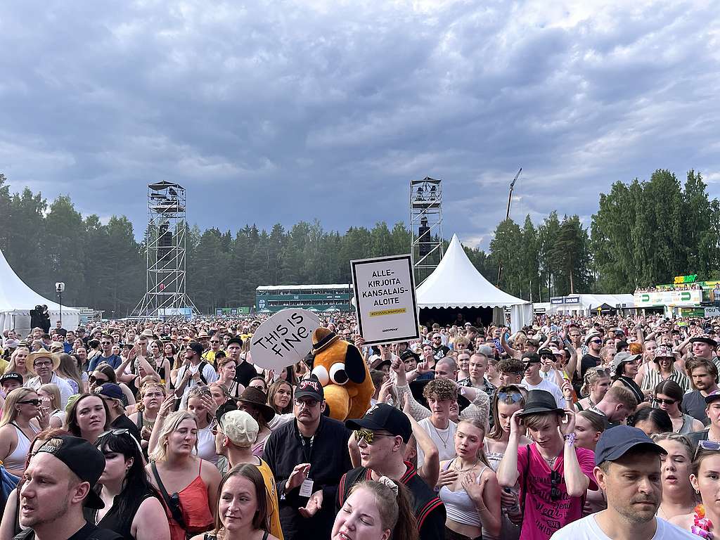 Festarikansaa provinssirockissa. Kuvan keskellä näkyy Greenpeacen kyltti "Allekirjoita kansalaisaloite" This is fine -meemikoiran vieressä.