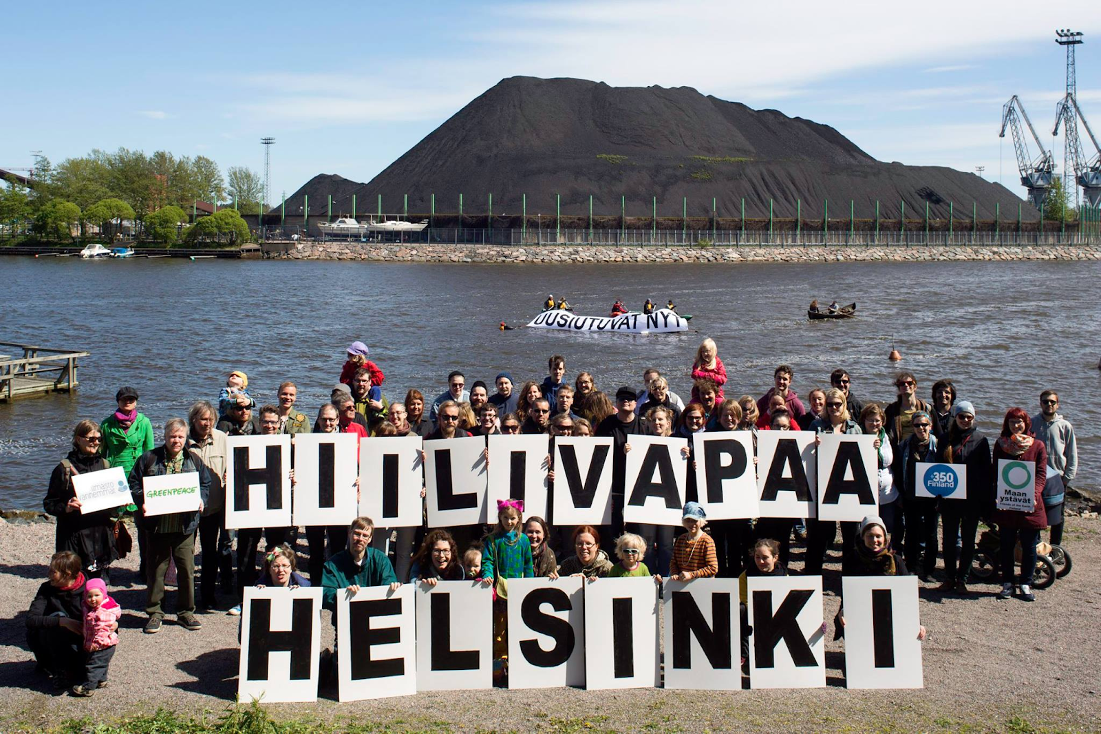 Mielenosoittajia Hanasaaren hiilivuoren edustalla. Mielenosoittajilla on kylttejä, joissa lukee mm. "uusiutuvat nyt" sekä "Hiilivapaa Helsinki".
