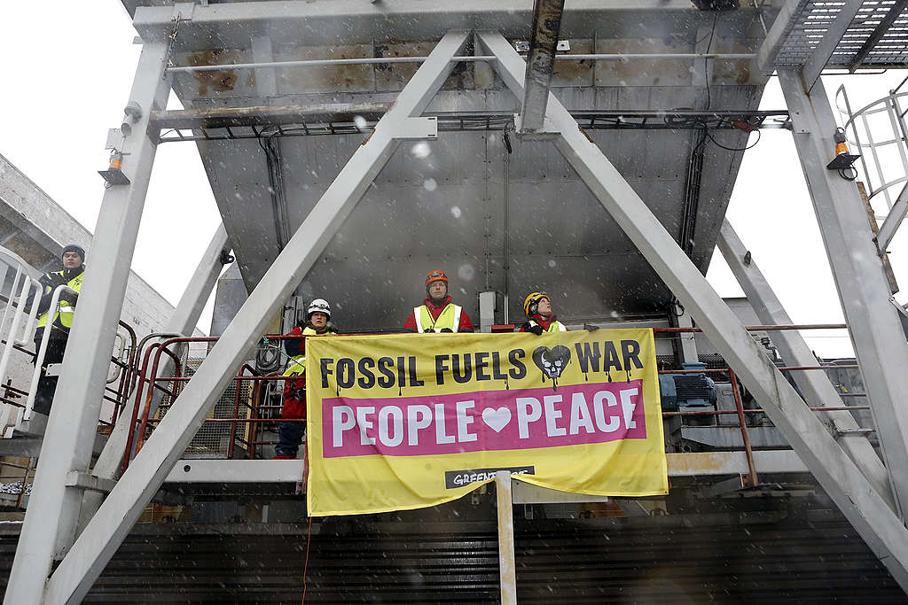Kolme aktivistia pitelee satamassa banderollia, jossa lukee "Fossil fuels love war, people love peace".